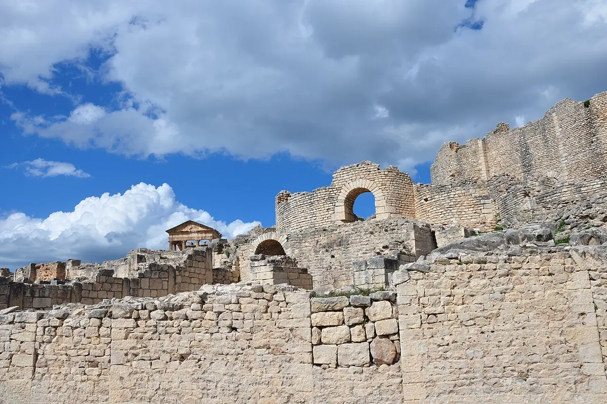 Pale limestone walls and arches of the Licinian Baths at Dougga, with a small temple front on the skyline beyond